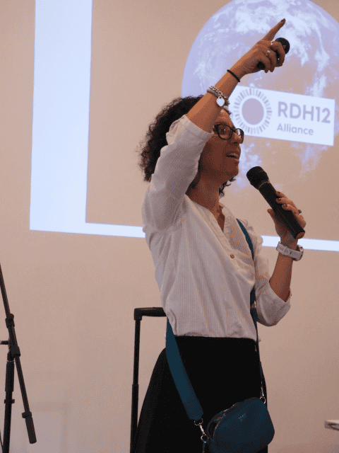 A person stands indoors holding a microphone and pointing upward while speaking, with a presentation slide projected behind them. The slide includes a globe graphic and the text “RDH12 Alliance.” The speaker wears a light shirt, dark trousers, and a crossbody bag.