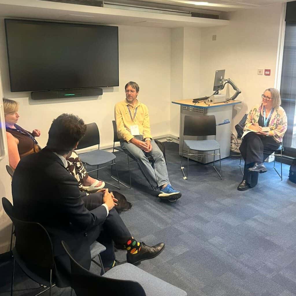 A small group of people seated in a circle of chairs in a meeting room, participating in a discussion. A large wall-mounted screen and a lectern with a computer are visible in the background.