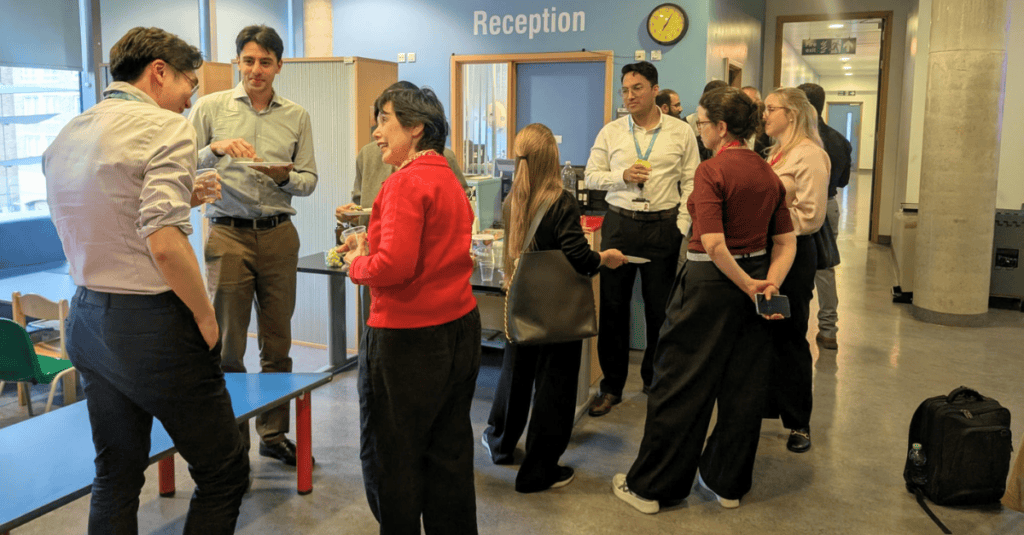 Colleagues in business-casual attire gather in a bright reception area, chatting around a table with refreshments.
A “Reception” sign and wall clock are visible, with bags and a suitcase placed along the side.