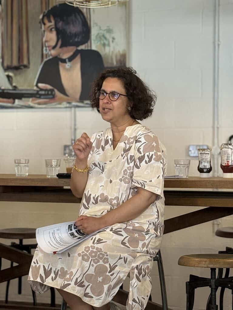 Prof Jugnoo Rahi is seated on a high stool speaking at a coffee hour event, holding printed notes. She is in a café with wooden tables, water glasses, and condiment bottles. A large piece of artwork featuring a person is visible on the wall behind.