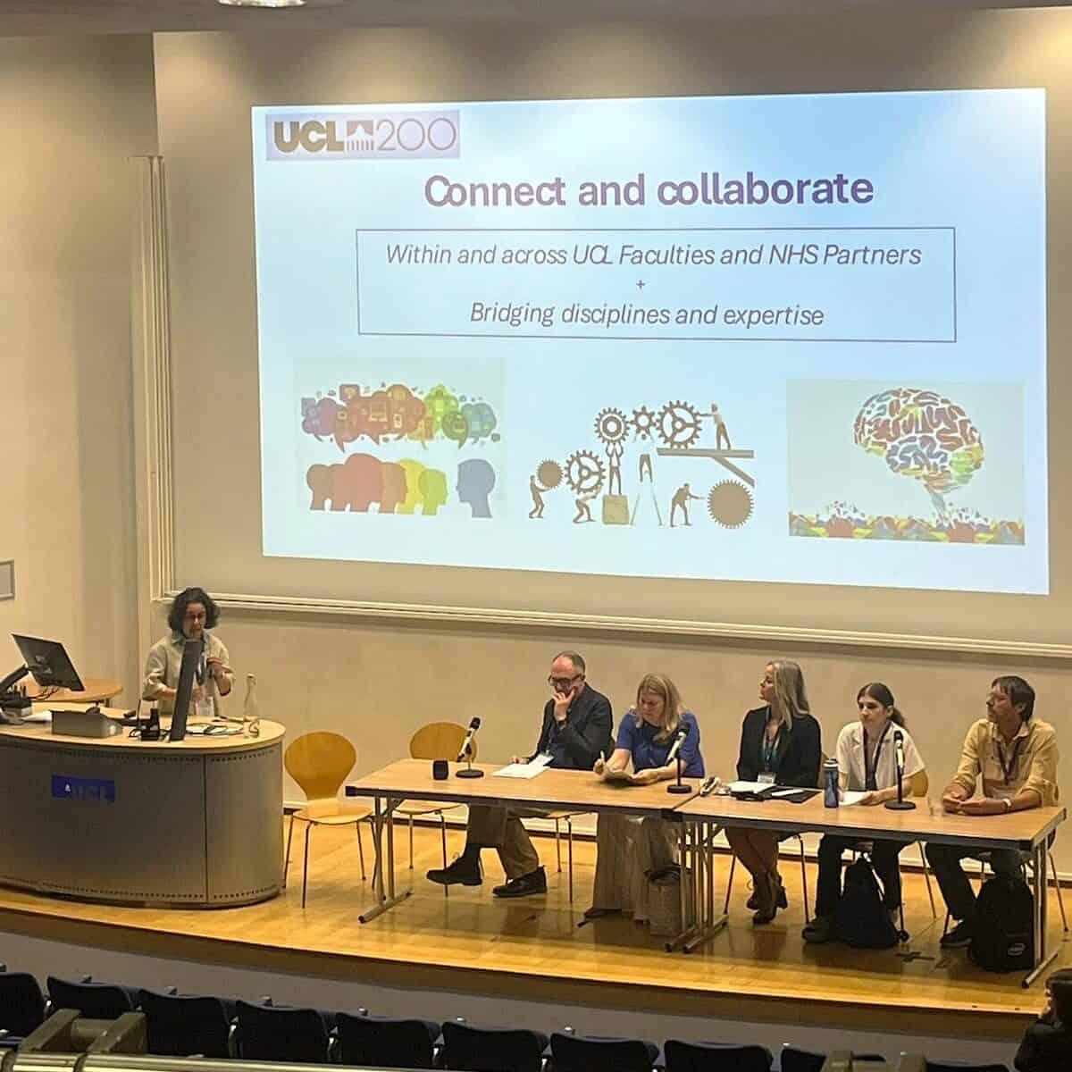 A panel session in a lecture theatre with several speakers seated behind a long table with microphones. Professor Jugnoo Rahi stands at a lectern. A large projected slide behind them reads “UCL200: Connect and collaborate”.