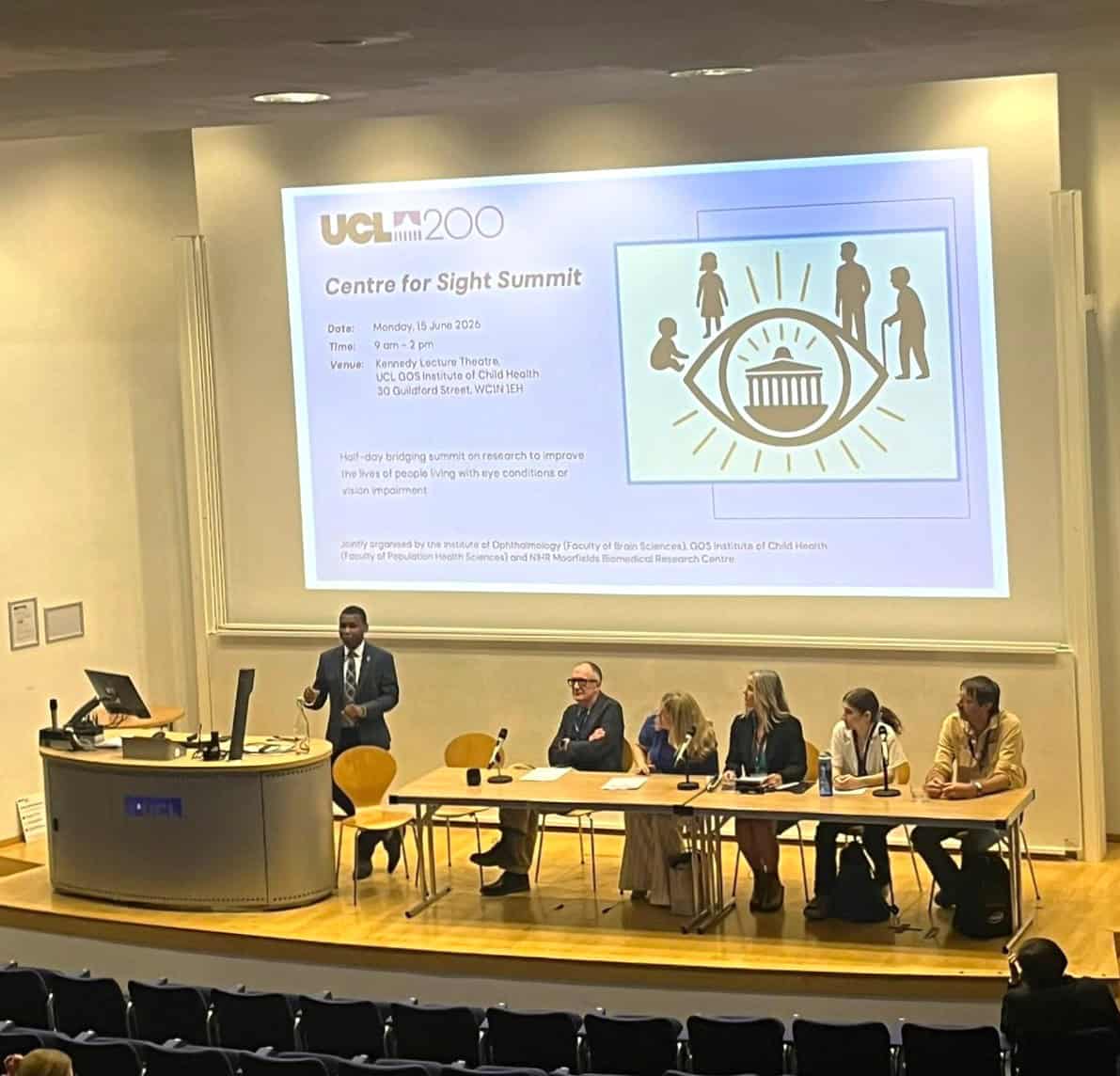 Professor Ibrahim Abubakar stands at a lectern addressing an audience while a panel of speakers sits at a long table on stage. A projected slide above them reads “UCL200 Centre for Sight Summit” and includes event details such as date, time and venue.