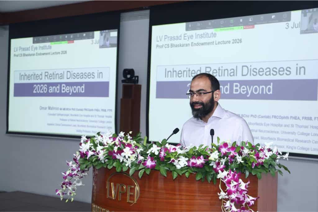 Prof. Mahroo wearing glasses and a light shirt stands at a flower-decorated podium, delivering a lecture in a conference room. Two large projection screens behind him display the presentation title: "Inherited Retinal Diseases in 2026 and Beyond." The header on the slides reads "LV Prasad Eye Institute" and "Prof CS Bhaskaran Endowment Lecture 2026." affiliations, including Moorfields Eye Hospital, University College London, and