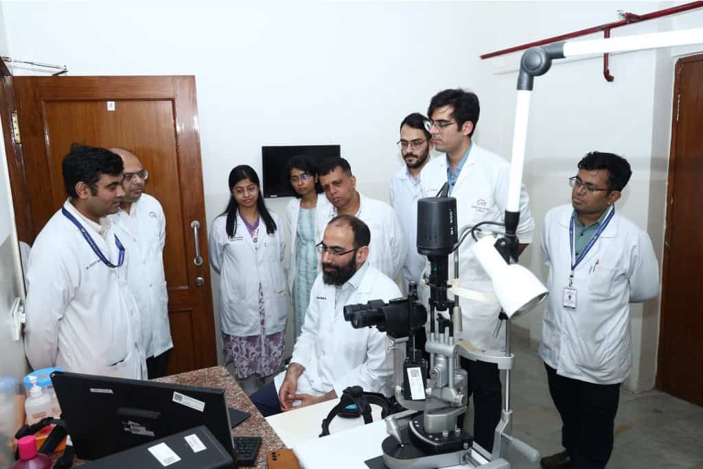 Image shows Prof. Mahroo in a clinical room surrounded by white coated clinicians. they are looking at what he is showing them on screen