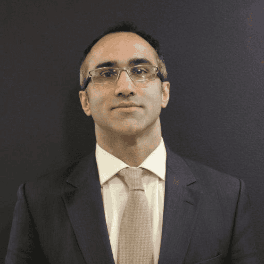 Professional head-and-shoulders portrait of a person wearing glasses, a dark suit jacket, white shirt and light-coloured tie, standing against a dark background and facing the camera.